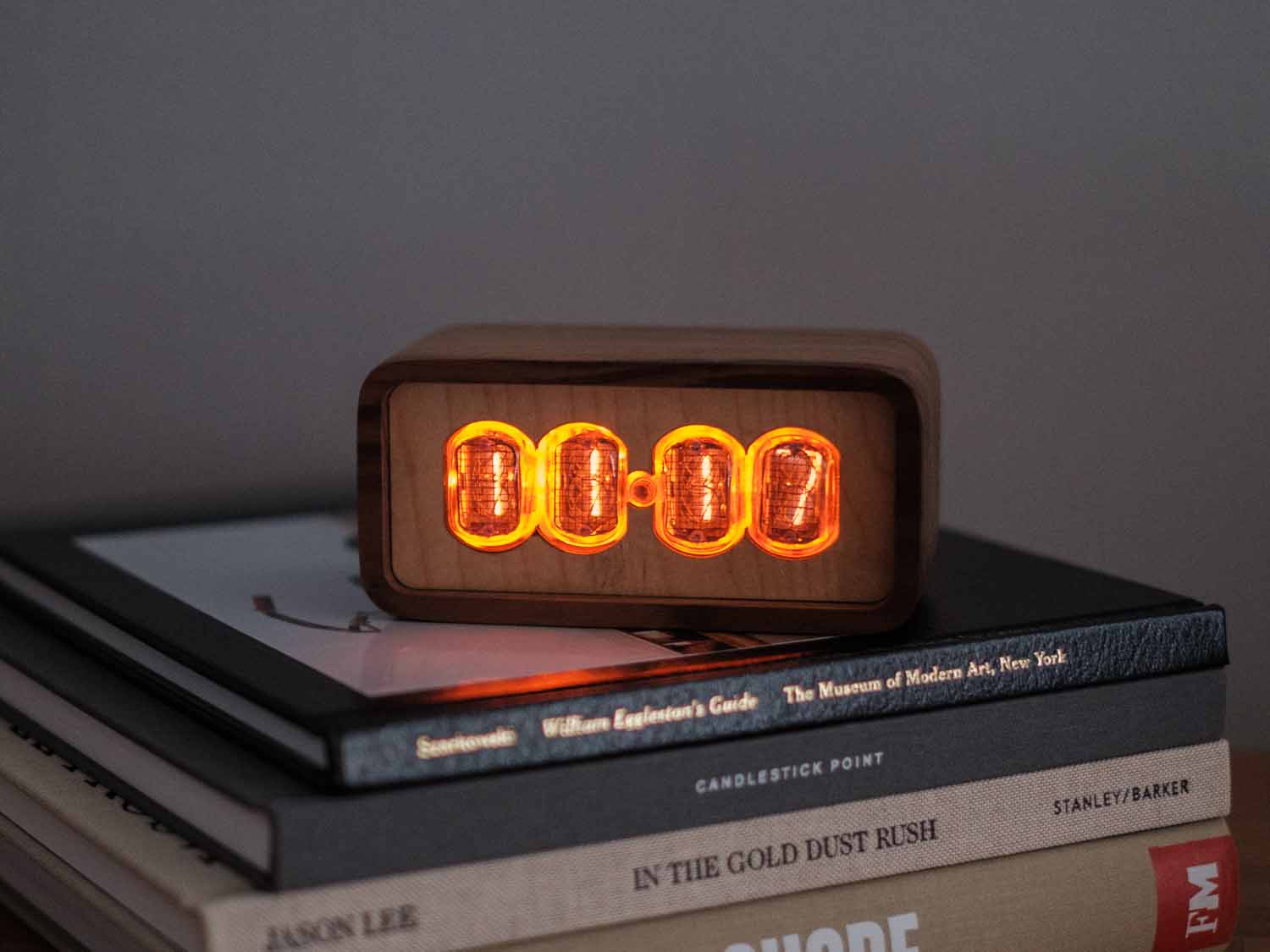 Clock sits on a stack of books and is made from solid walnut with a contrasting maple face. The design uses retro Nixie tubes for a warm orange glow.