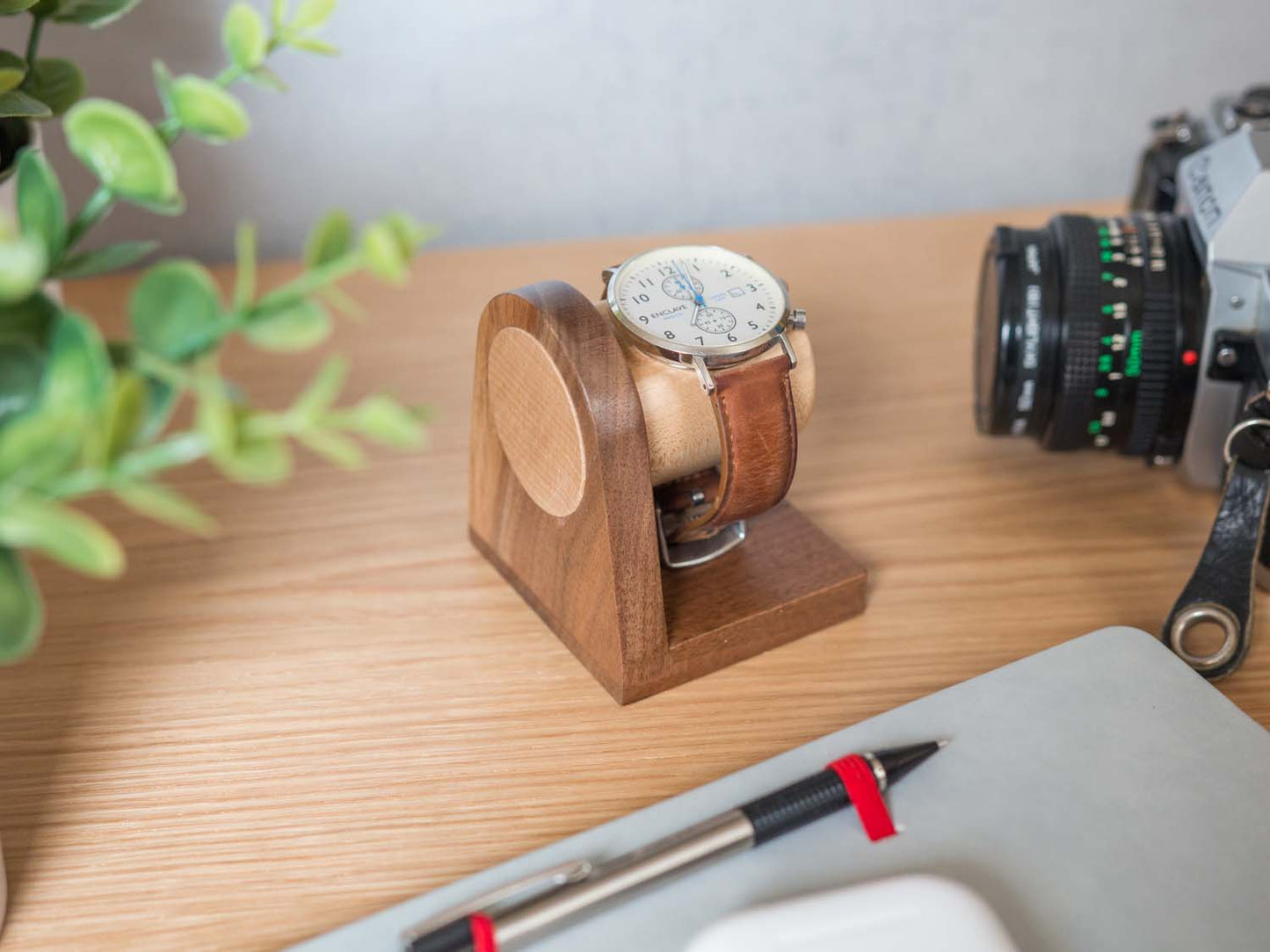 One watch rests on a smooth maple dowel which rests on a strong walnut base. Set on a desk with notebook and camera.