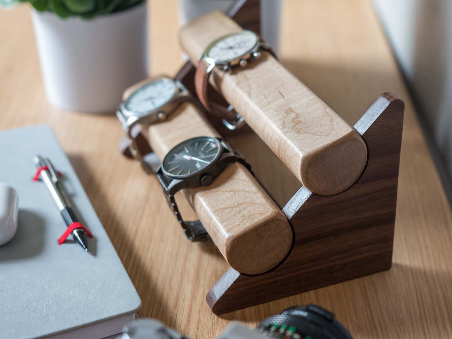 Three watches rest on a smooth maple dowel which rests on a strong walnut base. Set on a desk with notebook and camera. Warm wood nicely contrasts the mechanical metal watch.
