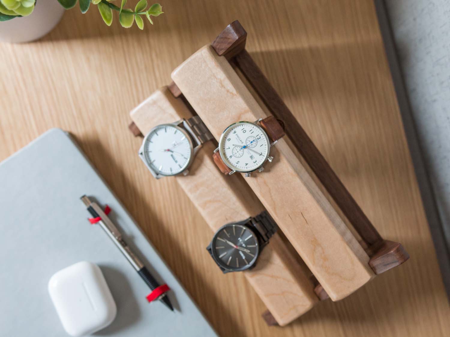 Three watches rest on a smooth maple dowel which rests on a strong walnut base. Set on a desk with notebook and camera. Warm wood nicely contrasts the mechanical metal watch.