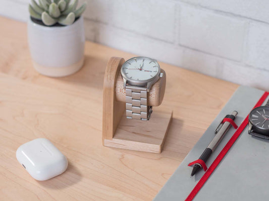 One watch rests on a smooth maple dowel which rests on a strong maple base. Set on a desk with notebook and camera. The warm wood contrasts nicely with the mechanical metal watch.