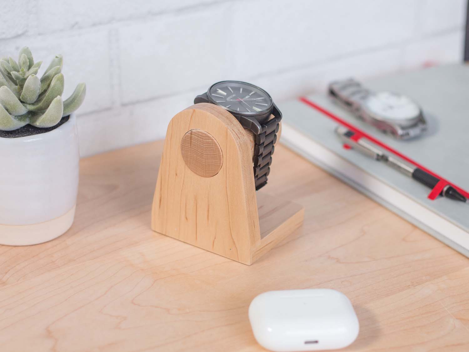 One watch rests on a smooth maple dowel which rests on a strong maple base. Set on a desk with notebook and camera. The warm wood contrasts nicely with the mechanical metal watch.