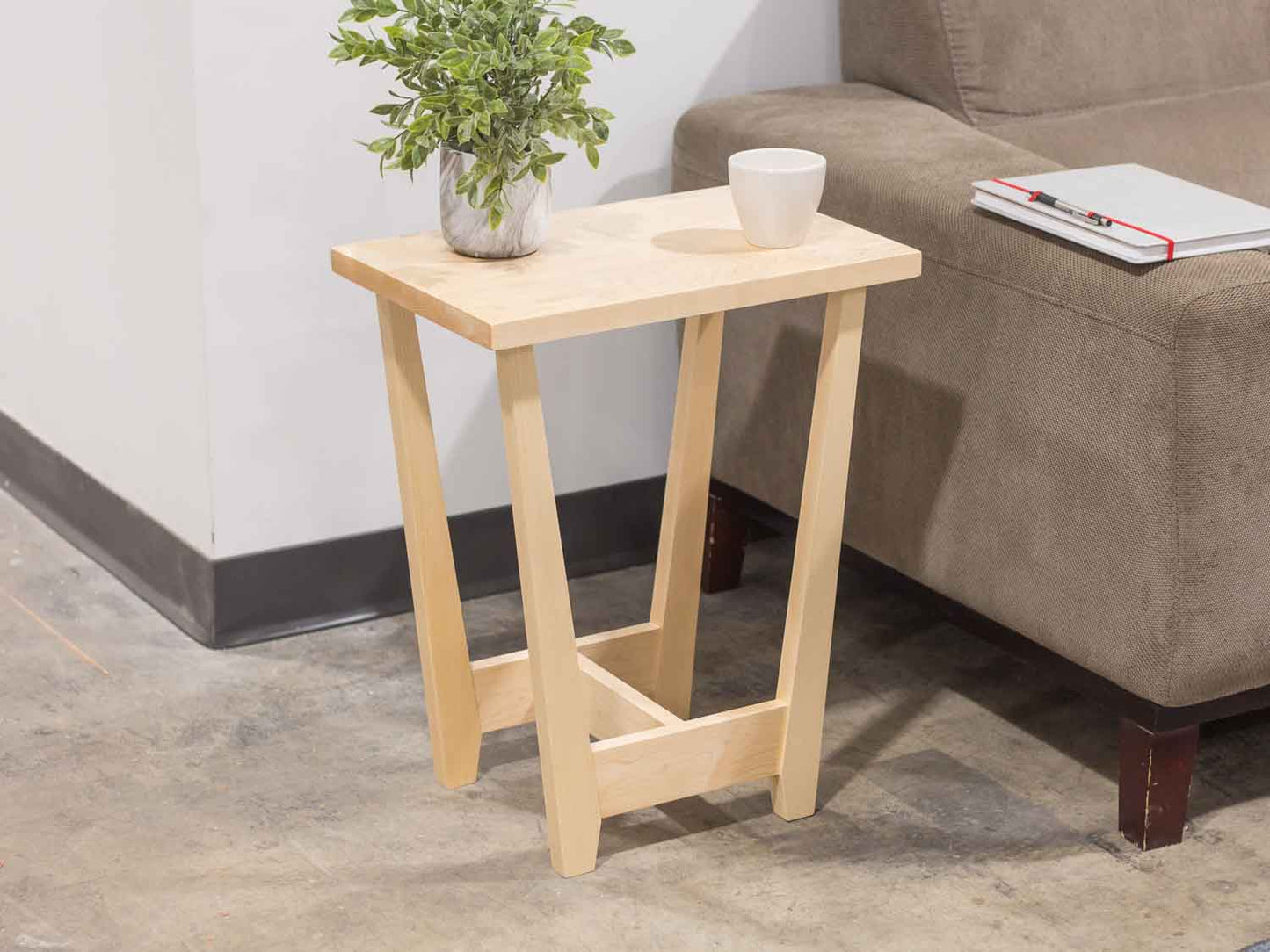 A side table placed in a living room with a plant and coffee mug sat on the table top. The side table is made from light maple hardwood and is crafted in Scandinavian minimalist modern design.