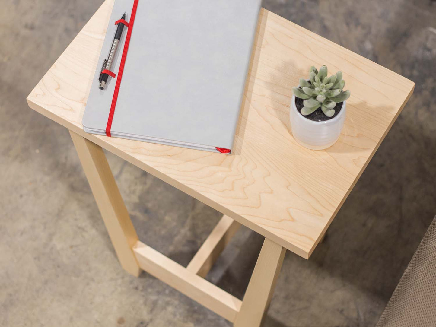 A side table placed in a living room with a plant and coffee mug sat on the table top. The side table is made from light maple hardwood and is crafted in Scandinavian minimalist modern design.