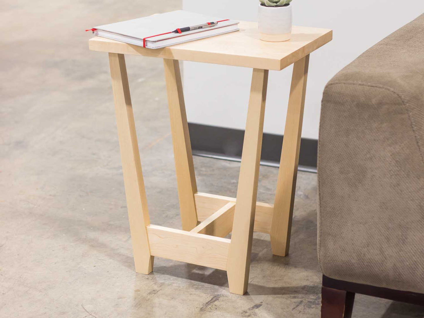 A side table placed in a living room with a plant and coffee mug sat on the table top. The side table is made from light maple hardwood and is crafted in Scandinavian minimalist modern design.